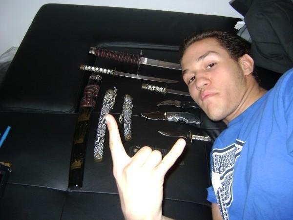 A young man lying on a black surface, making a hand gesture, with two knives and various sword-like objects arranged behind him.