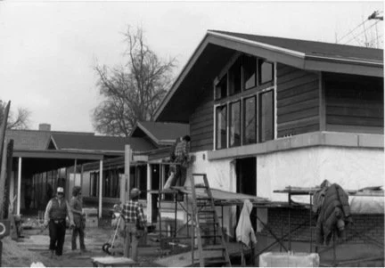 Construction workers building a house, with some on scaffolding, in a suburban neighborhood.