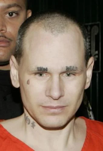 Close-up of a man with a bald head, tattoos on his neck, wearing a red shirt, and another person partially visible behind him.