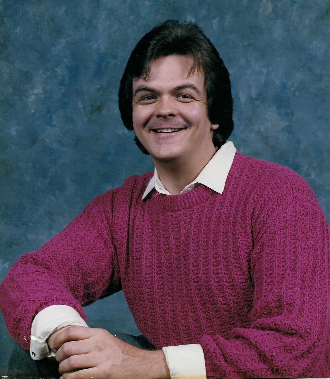 A young man with dark hair and a big smile, wearing a burgundy knit sweater over a white collared shirt, is sitting with his hands clasped on a table against a mottled blue and green backdrop.