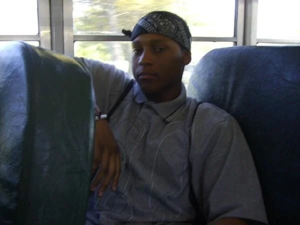 A young man sitting on a bus seat, wearing a gray shirt and a patterned bandana, with trees visible through the window behind him.
