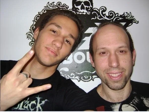 Two men taking a selfie in front of a black and white wall art featuring a skull and intricate designs.