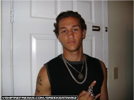 Young man with curly hair, wearing a black sleeveless shirt and jewelry, standing in front of a white door, making a hand gesture.