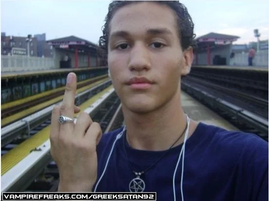 Young man taking a selfie at a train station, pointing one finger up, wearing a dark shirt, with earphones, a ring, and a pentagram necklace.