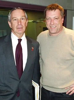 Two men standing together, one in a suit and tie and the other in a casual sweater, smiling for the camera.