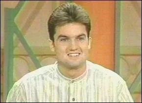 A young man with brown hair smiling and wearing a light-colored striped shirt, standing in front of a colorful background.
