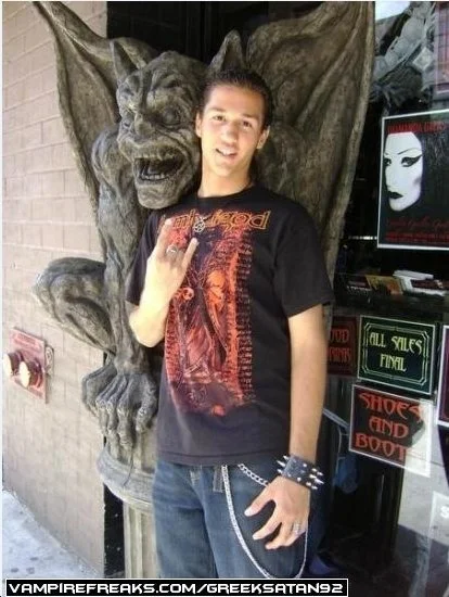 A young man standing in front of a decorative gargoyle sculpture outside a building, giving the rock on sign with his hand, and wearing a black graphic t-shirt and studded bracelets.