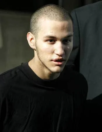 A young man with a shaved head wearing a black shirt, looking slightly to the side indoors.