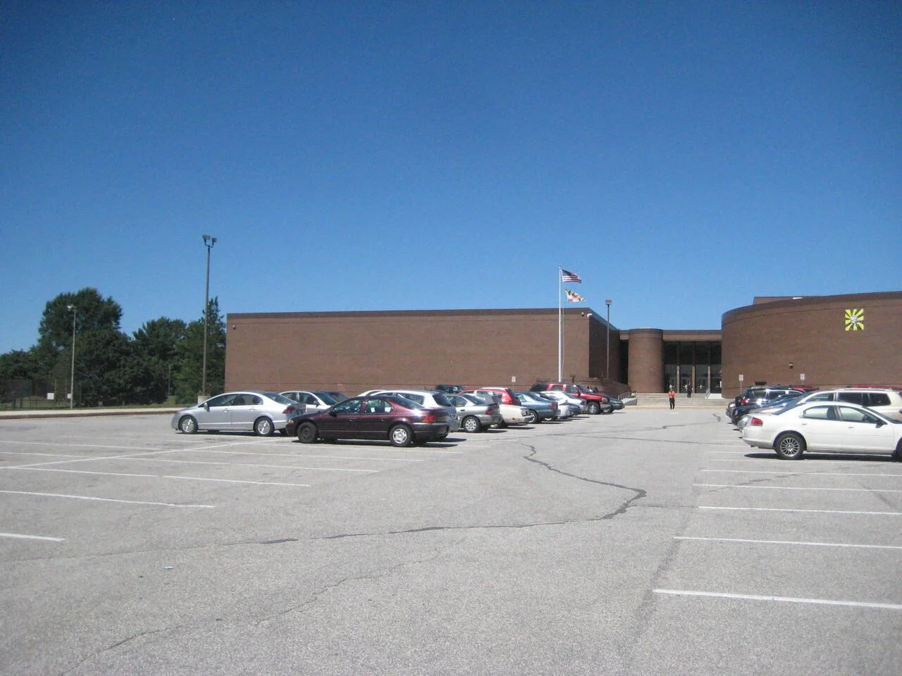 View of a large parking lot in front of a brick building with flags, including the American flag, flying on a flagpole. Cars are parked in the lot, and a few people are walking toward the entrance.