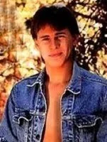 A young man with dark hair wearing a denim jacket, standing outdoors with trees and golden leaves in the background.