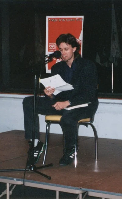 Young man reading from paper at microphone on stage, seated on a chair with a yellow cushion, in a room with black curtains and a red poster in the background.