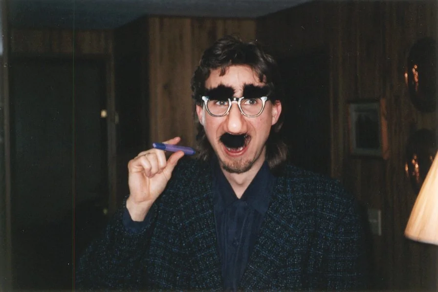 Person with exaggerated eyebrows, mustache, and glasses, holding a pen, indoors with wood-paneled walls.