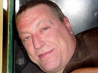 A close-up of a man smiling, with short hair and wearing a brown shirt, next to a green object.