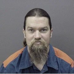 Portrait of a man with long dark hair tied back, a full beard and mustache, wearing a blue and orange jacket against a gray background.