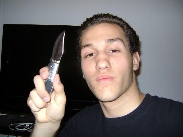 A young man with dark hair holding a large kitchen knife in his right hand, looking at the camera with a serious expression, standing in front of a black screen or monitor.