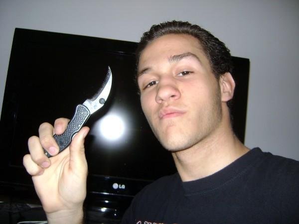 Young man with slicked-back hair holding a folding knife in front of a flat-screen TV.