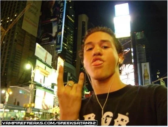 Young man taking a selfie in Times Square at night, with bright digital billboards and city buildings in the background, making a rock hand gesture.