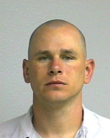 Headshot of a young man with a shaved head, wearing a white shirt, standing against a plain light-colored wall.