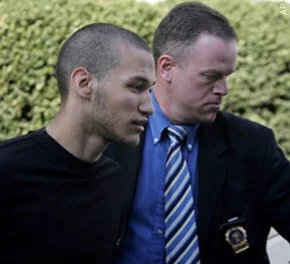 Two men with serious expressions looking down, one in casual clothing and the other in a suit and tie, outdoors with greenery in the background.