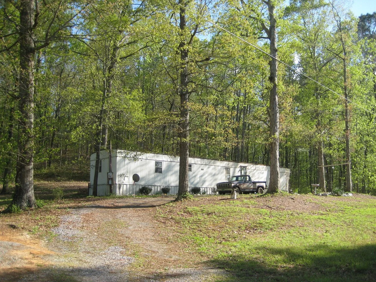 A mobile home in a wooded area with trees and green grass, a gravel driveway, and an old pickup truck parked near the mobile home.