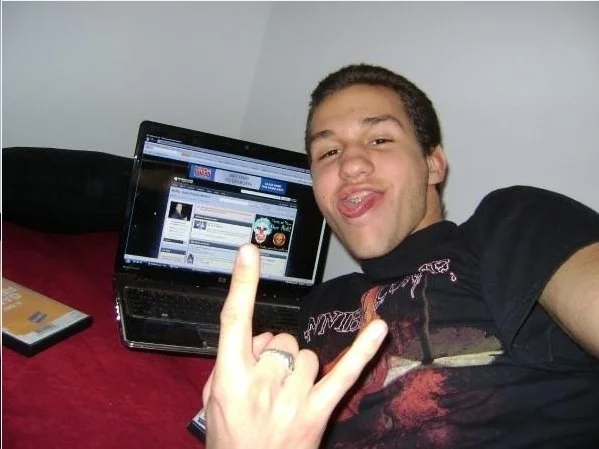 Young man lying in bed, taking a selfie while using a laptop on his lap. He is making a rock and roll hand gesture with his right hand and smiling.