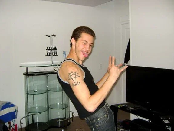 A young man with short dark hair, wearing a black shirt with white trim and jeans, making a playful face while holding a remote control in his right hand and showing a peace sign with his left hand. He is standing in a living room near a flat-screen TV.