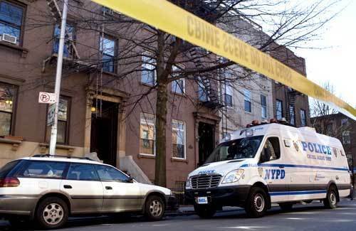 A police van and a parked car behind police crime scene tape on a city street with brown brick buildings.