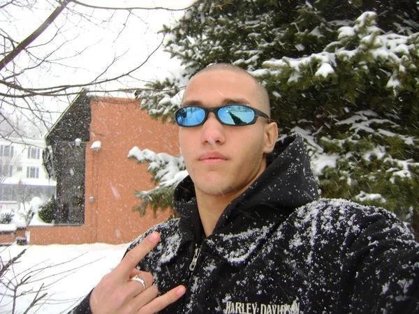 A young man with short hair wearing reflective sunglasses and a black Harley-Davidson hoodie, taking a selfie outdoors in a snowy winter landscape.