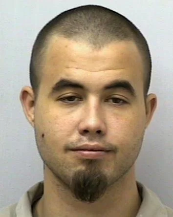 Close-up of a young man with a short buzz cut hairstyle, light facial hair, and a neutral expression, wearing a beige shirt against a plain gray background.