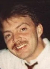 A man with brown hair and a mustache, smiling, wearing a white shirt with a dark tie.