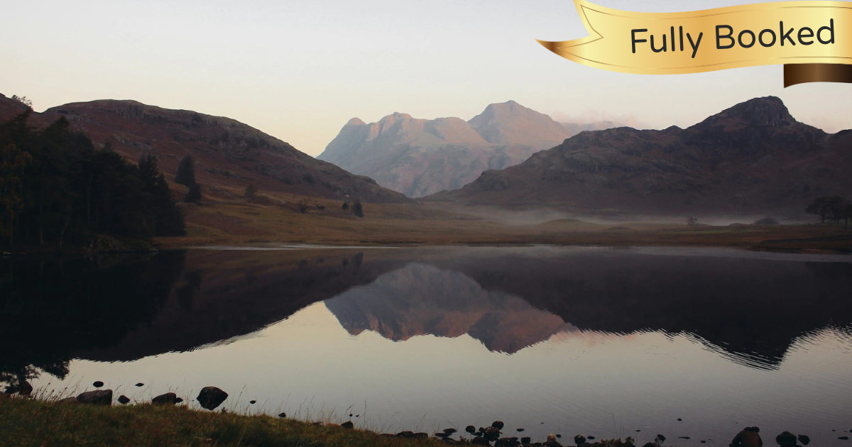 A serene landscape of mountains reflected in a calm lake during dusk or dawn, with a grassy shoreline and rocks in the foreground.
