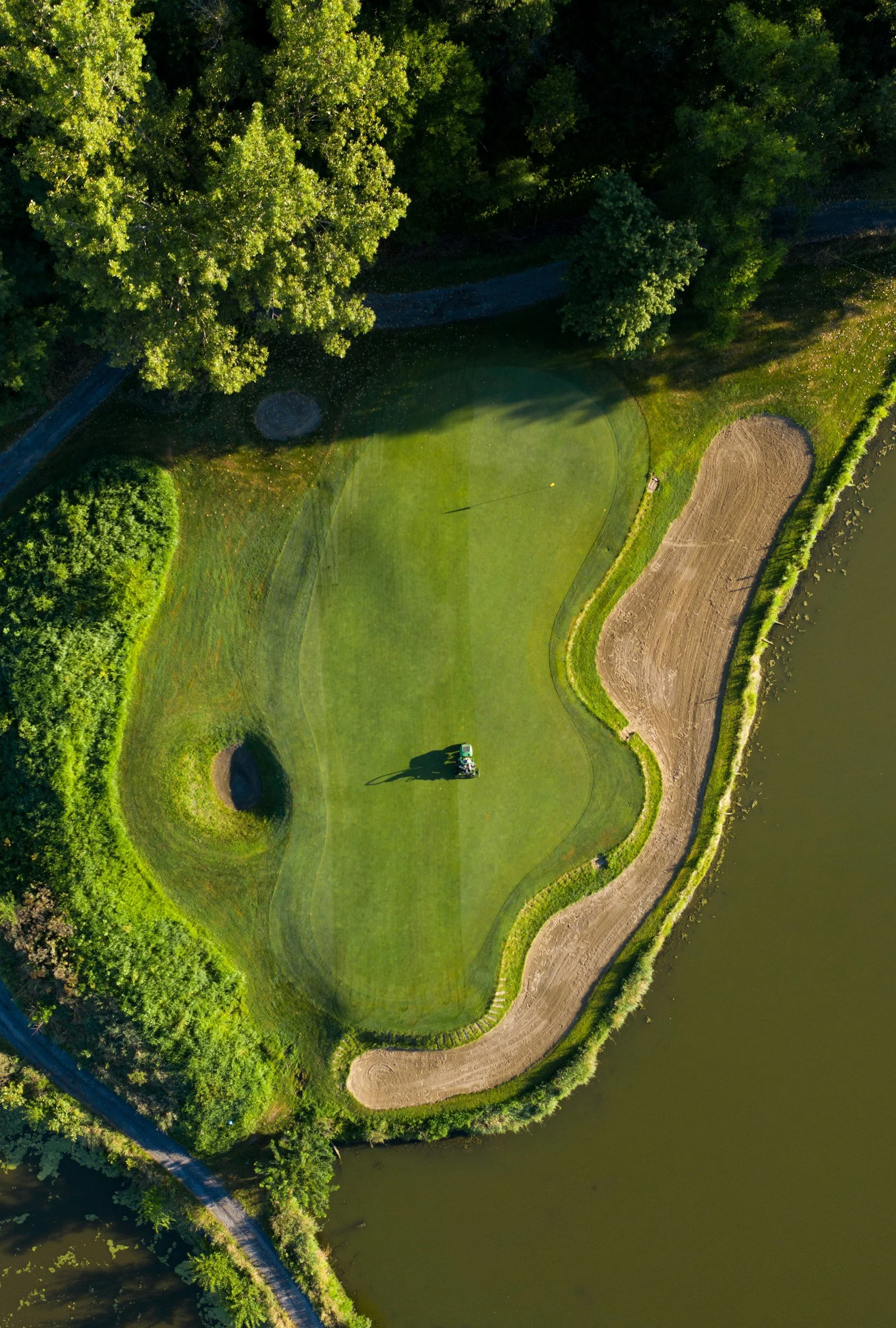 Aerial view of a golf course green surrounded by trees, water, and sand traps with a golf cart on the fairway.