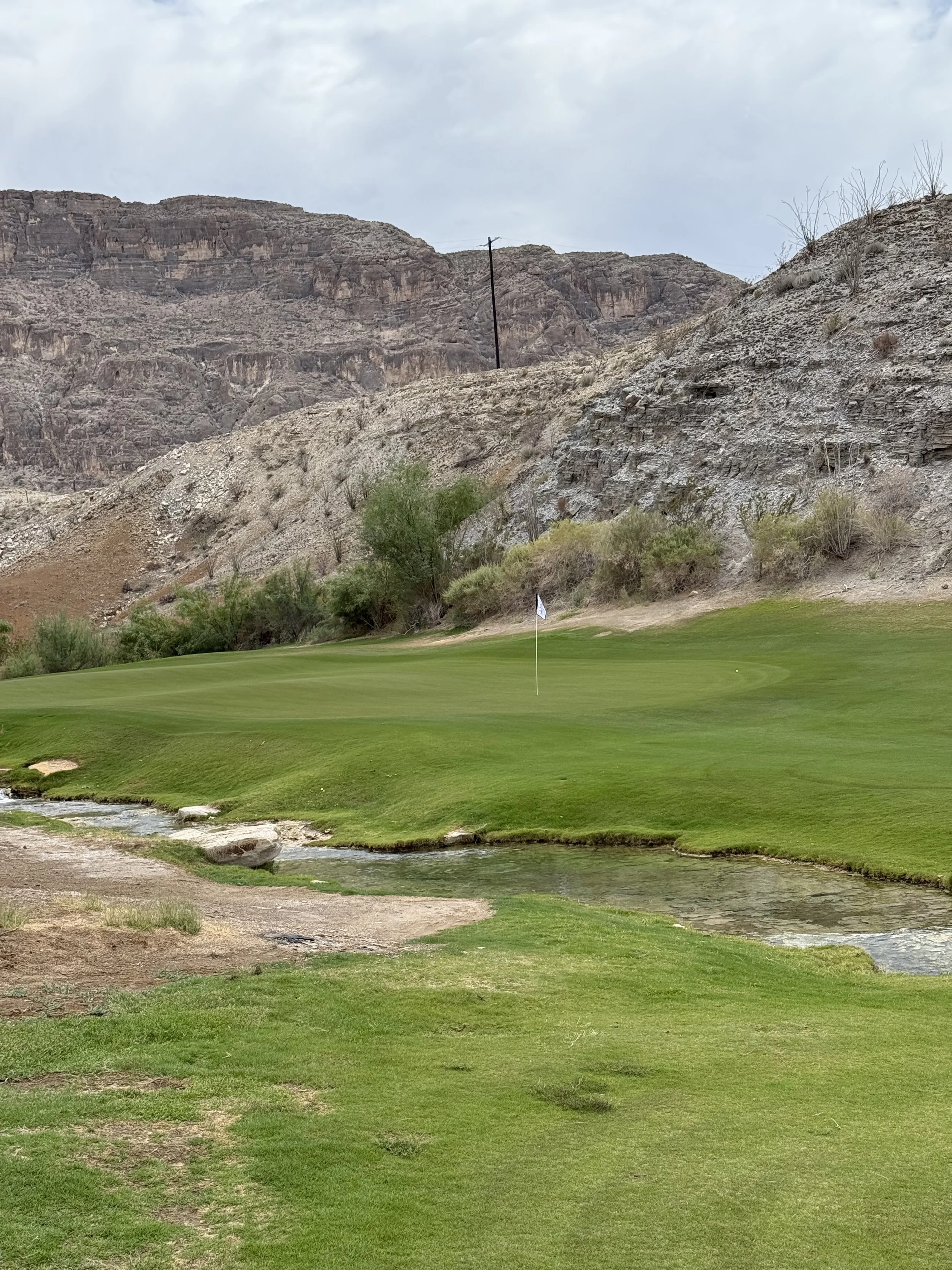 A golf course with green grass, a small water hazard, a flag on the putting green, and rocky hills in the background.