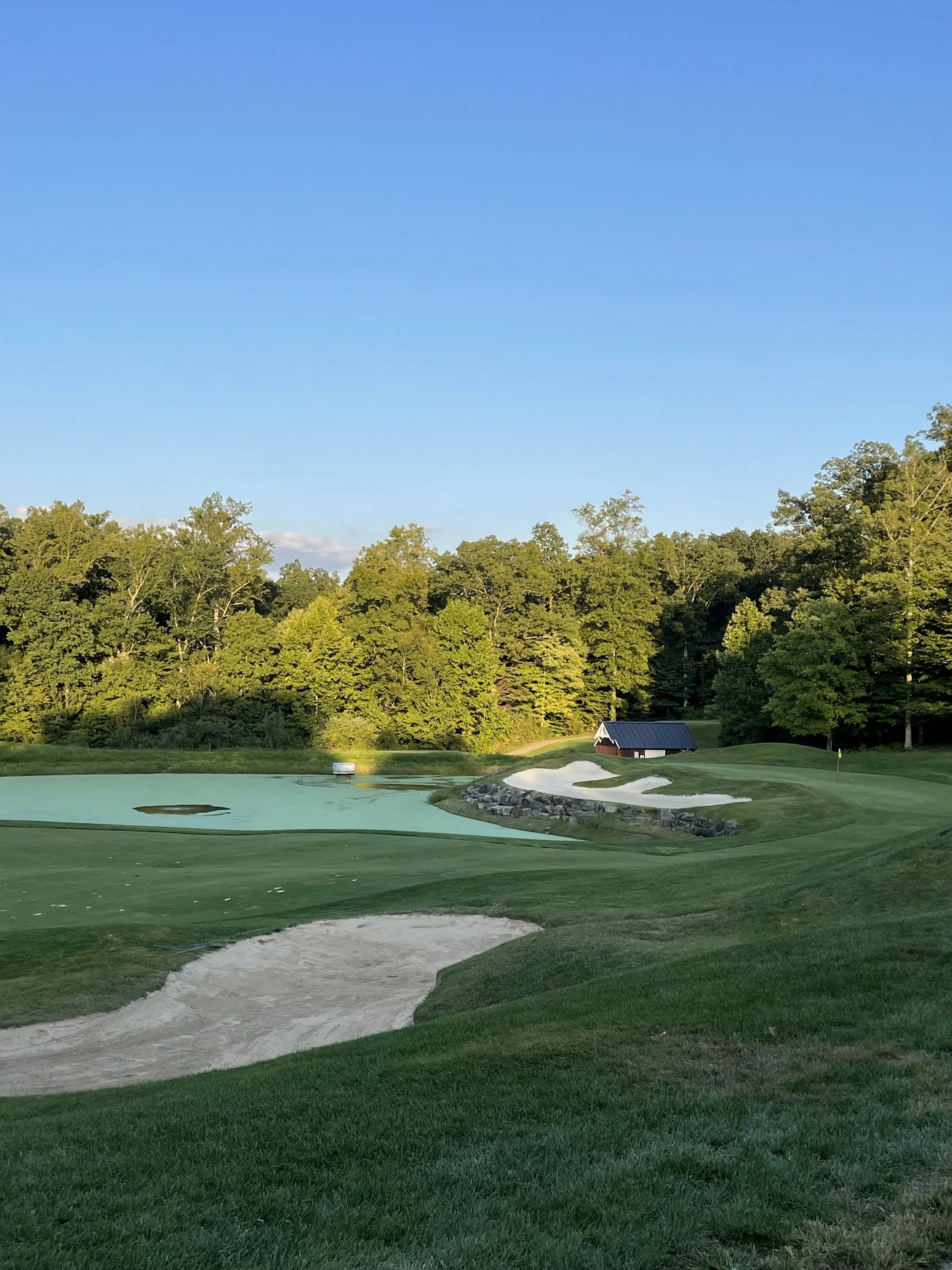 Scenic golf course with sand traps, a green, a water hazard, a small shed, and a backdrop of trees under a clear blue sky.