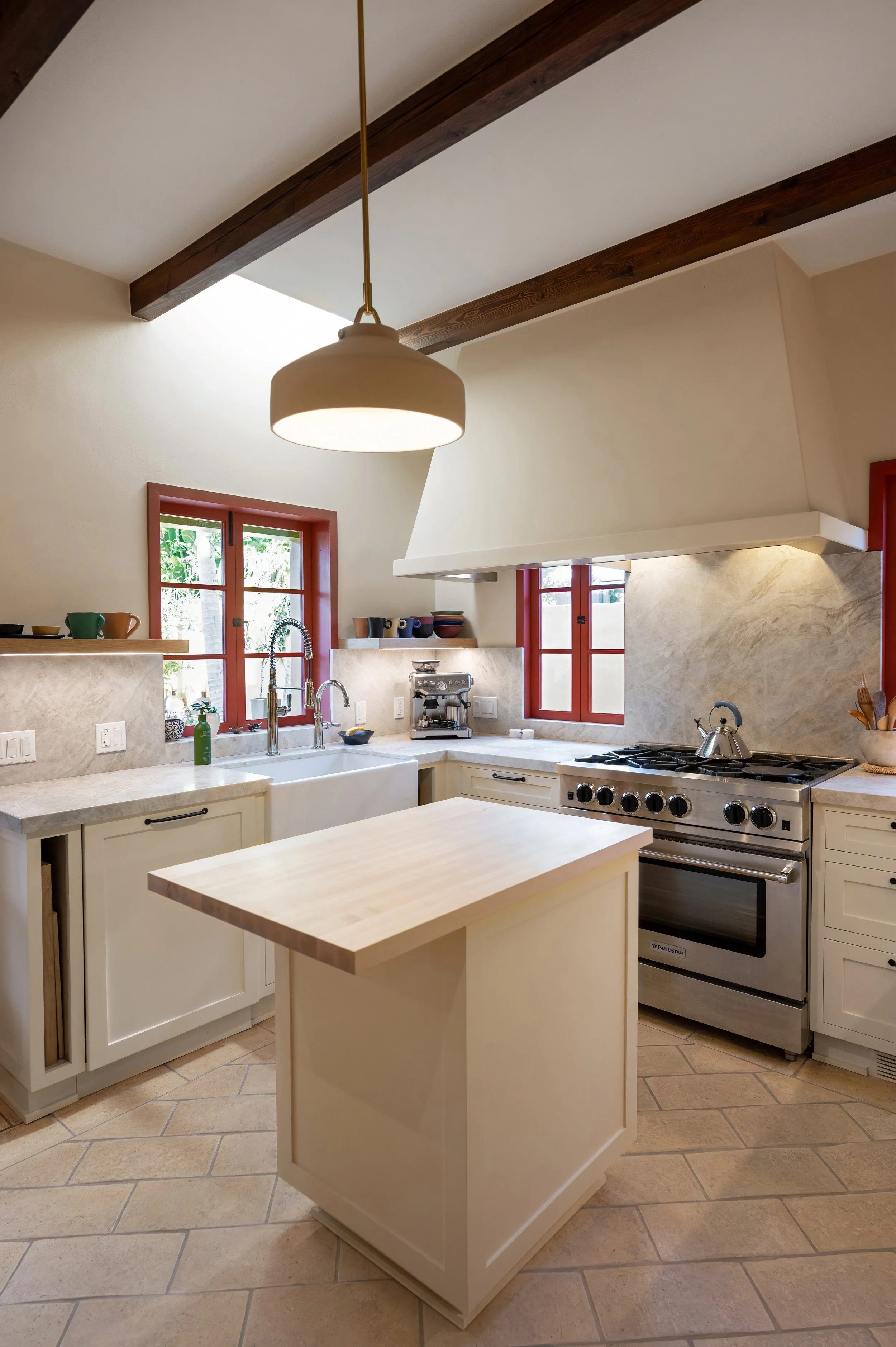 A kitchen with beige cabinets, a small kitchen island, a stainless steel stove, a white farmhouse sink beneath two red-framed windows, and a beige countertop. There is a hanging light fixture, and wooden beams on the ceiling.