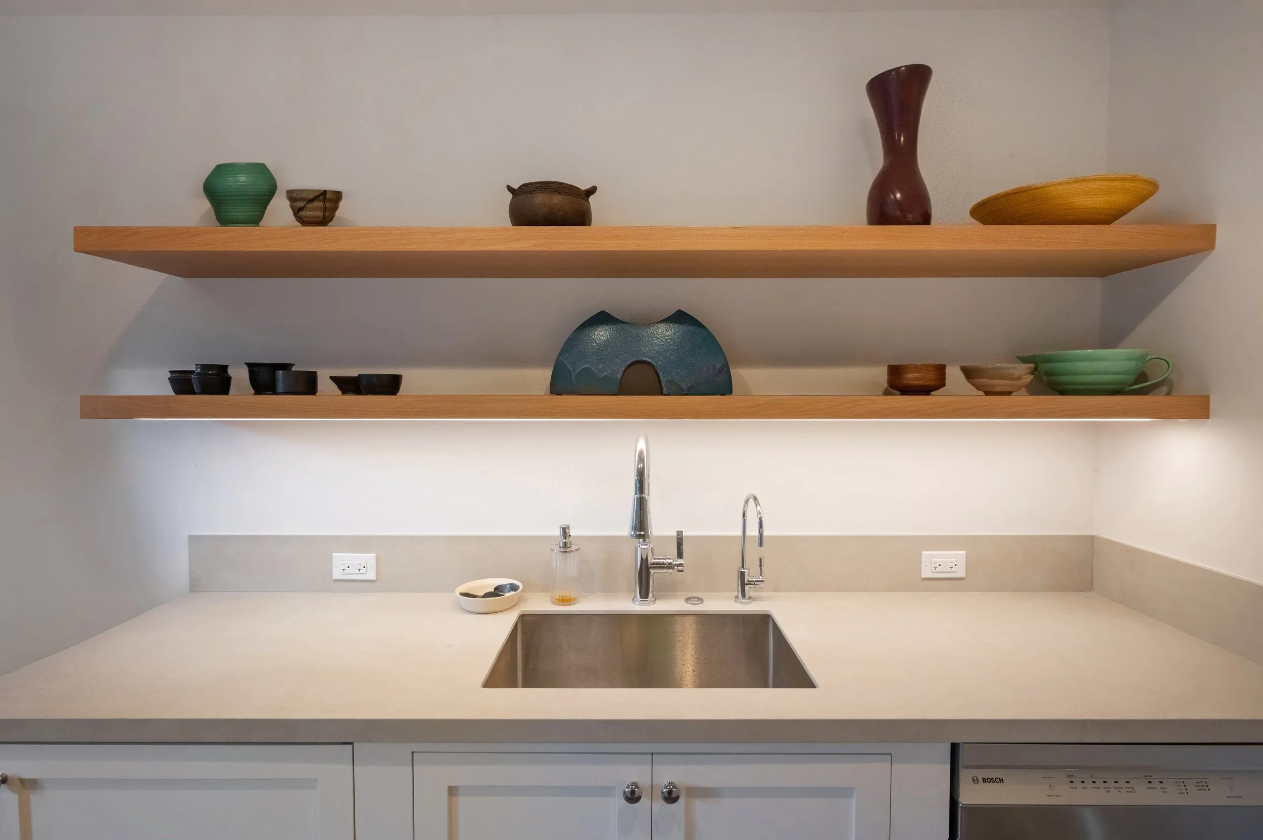Kitchen with open wooden shelves holding decorative pottery, a sink with a modern faucet, and a dishwasher beneath the counter.