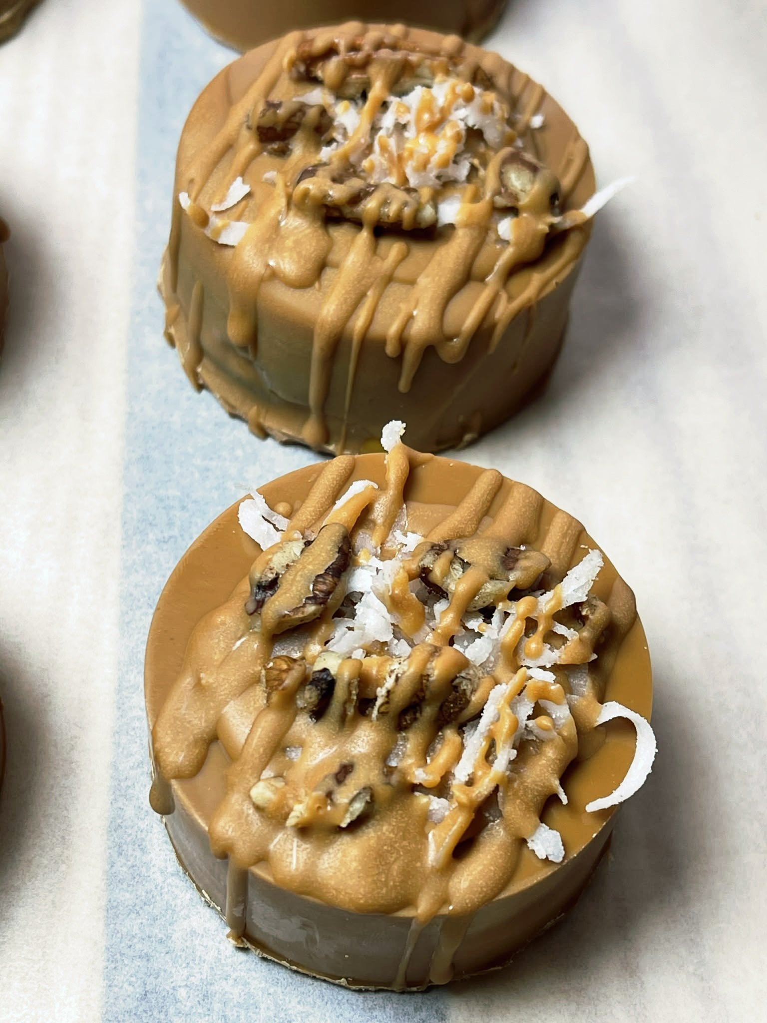 Two round caramel-flavored cake pucks topped with drizzled caramel sauce, shredded coconut, and chopped nuts.