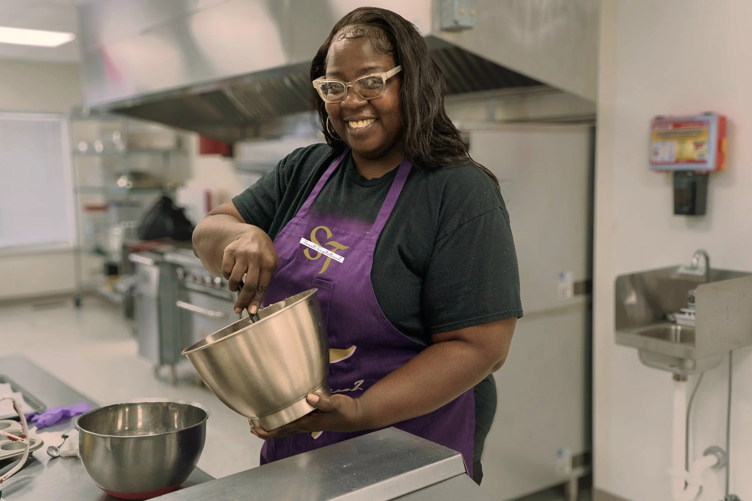 The CEO of Sweet TemptationZ with glasses and a purple apron smiling while mixing ingredients in a stainless steel bowl in a commercial kitchen.