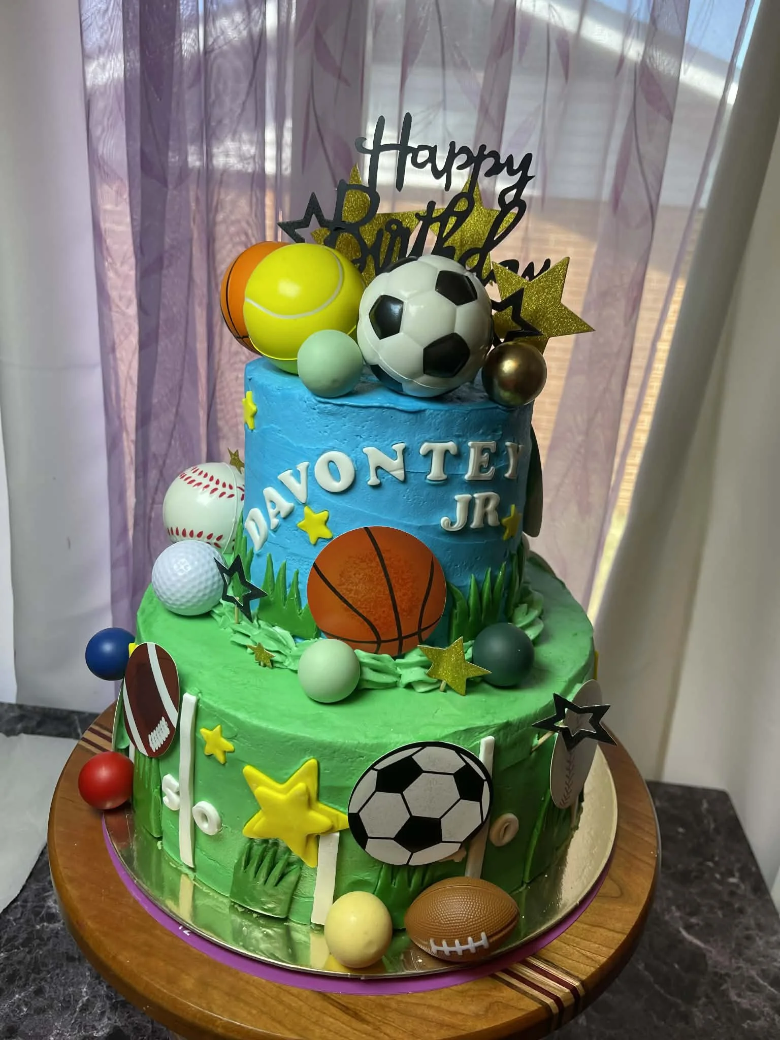 Two-tiered birthday cake decorated with sports-themed edible and non-edible decorations, including soccer balls, footballs, baseballs, basketball, star-shaped confetti, and a gold 'Happy Birthday' topper, with the name 'Davonte Jr.' on the front.