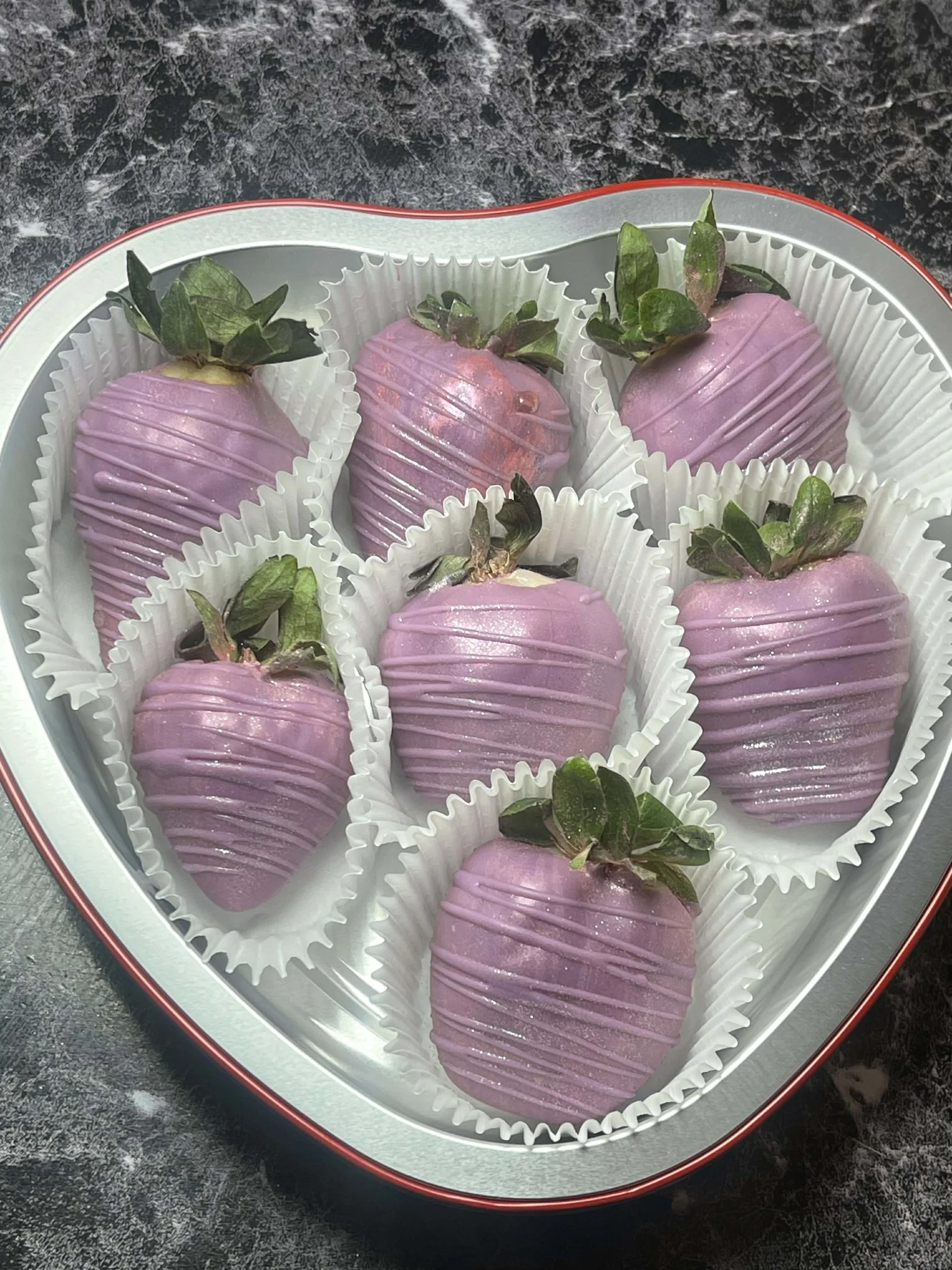 A heart-shaped box contains eight strawberries dipped in pink chocolate, decorated with white drizzle, each placed in a white paper cup.