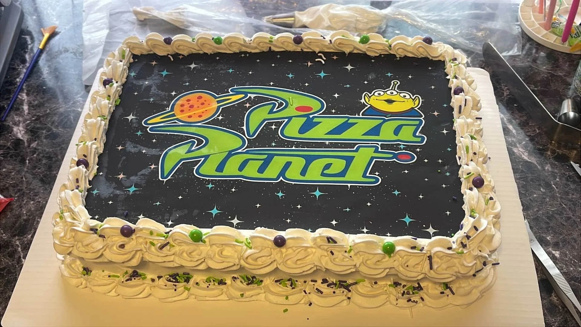 A rectangular birthday cake with a space-themed design featuring the words "Buzz Planet" and a cartoon alien, decorated with white frosting, colorful sprinkles, and purple and green candies.