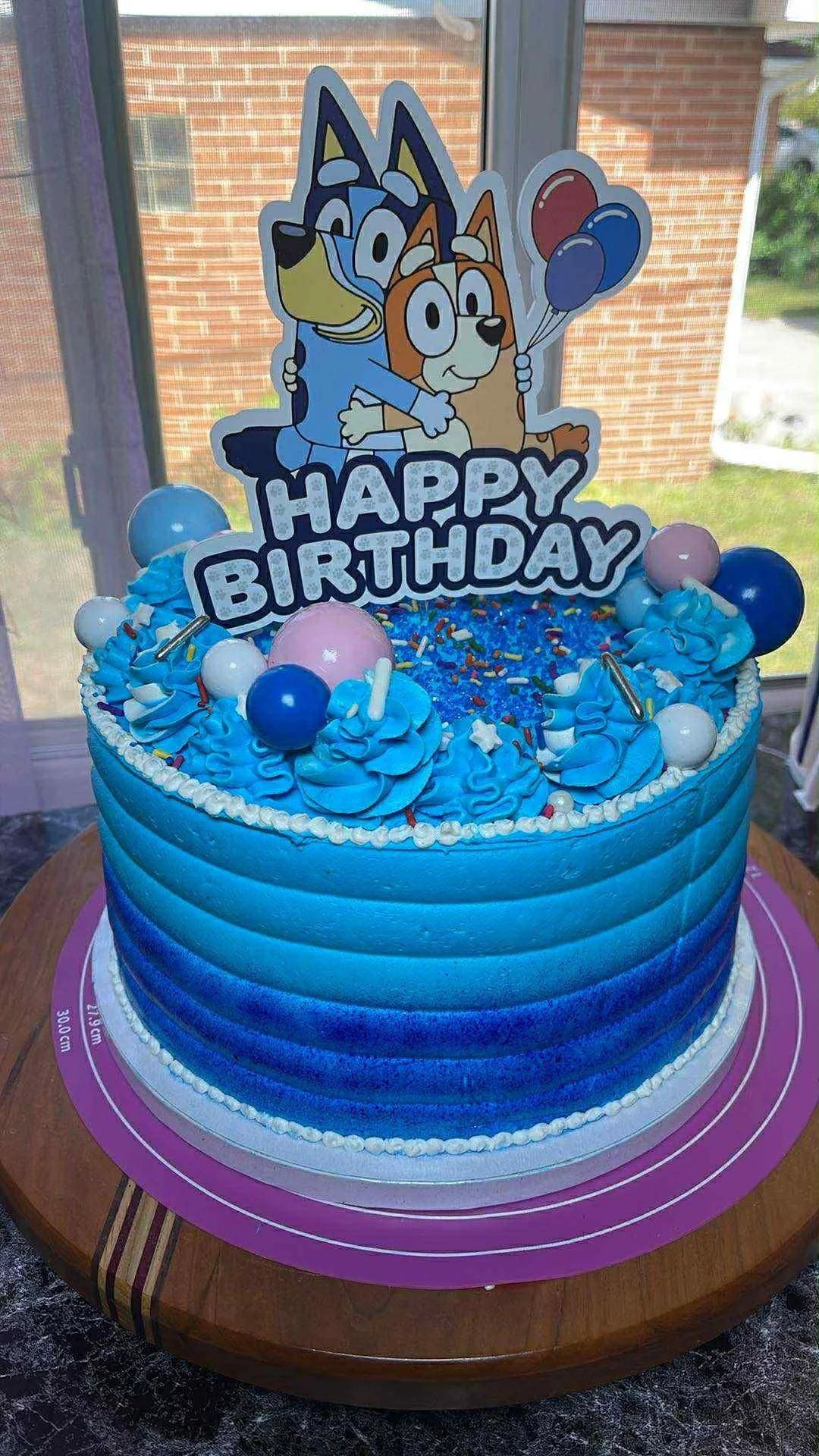 A birthday cake with blue gradient frosting, decorated with balloons and sprinkles. A cartoon cake topper features two dogs hugging, with a 'Happy Birthday' sign, one holding balloons. The cake is on a wooden table near a window.