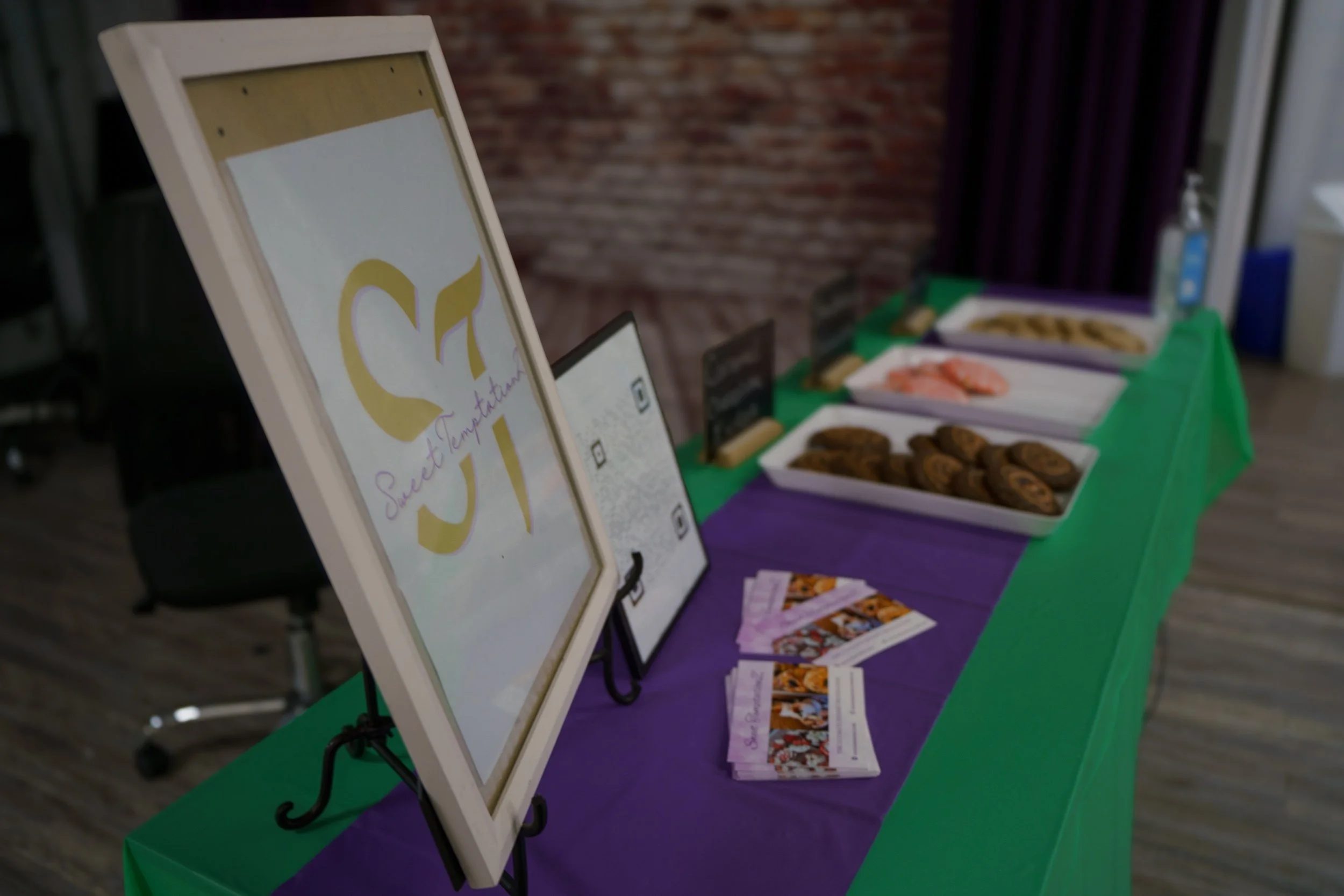 Display table with assorted cookies and promotional flyers at a bakery or event booth, with a sign that reads 'Sweet Temptations'.