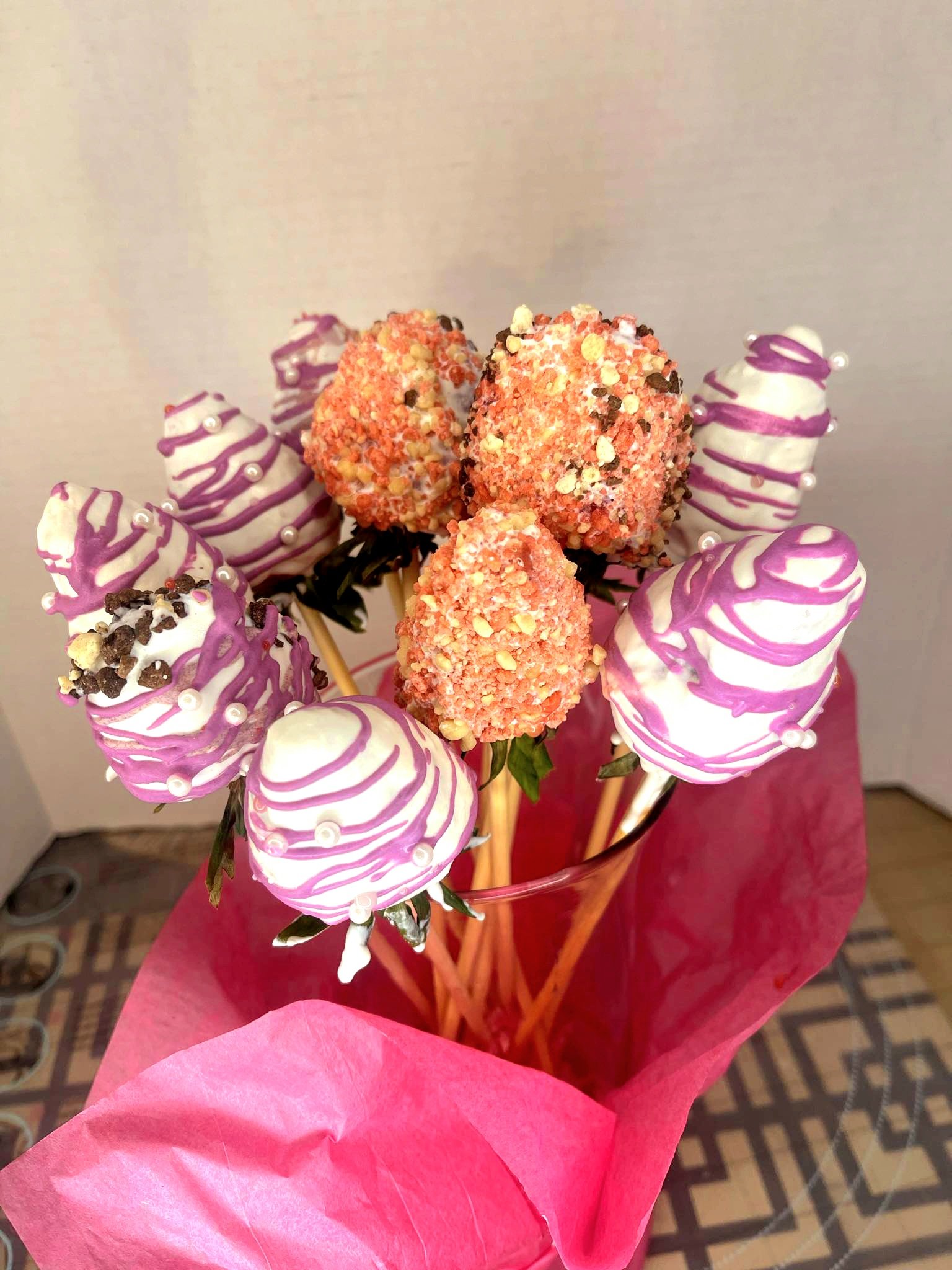 A bouquet of cake pops decorated with pink, white, and chocolate coatings, arranged in a glass vase wrapped with pink tissue paper.
