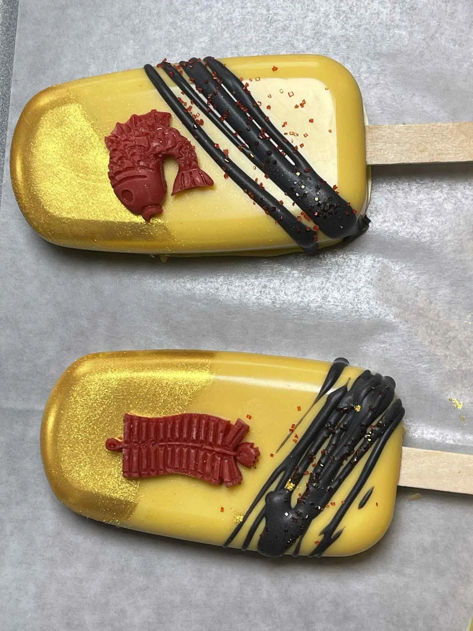 Two yellow and black popsicles decorated with red and black toppings and candies, placed on a gray surface.