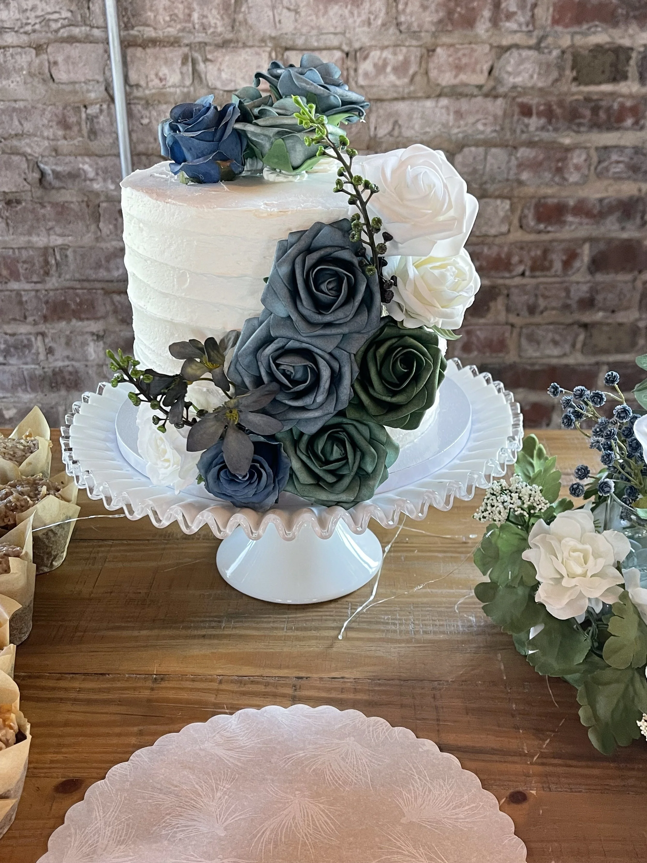 White tiered cake decorated with blue, gray, green, and white roses on a glass cake stand, with a bouquet of mixed flowers to the right, on a wooden table against a brick wall.