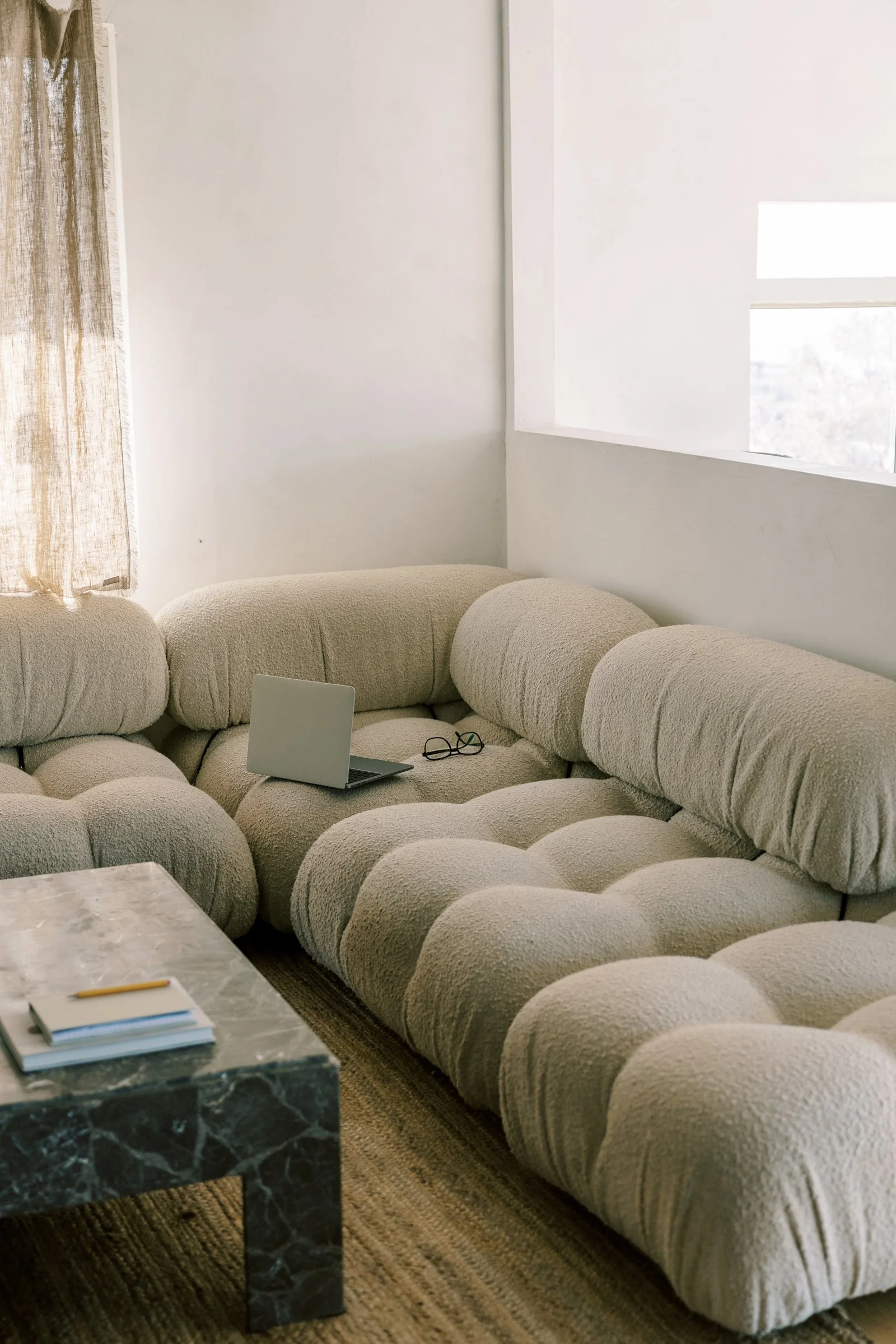 Living room with a beige, plush, sectional sofa, a marble coffee table with notebooks and a pencil, a laptop, and glasses on the sofa, and a window with a sheer curtain letting in natural light.