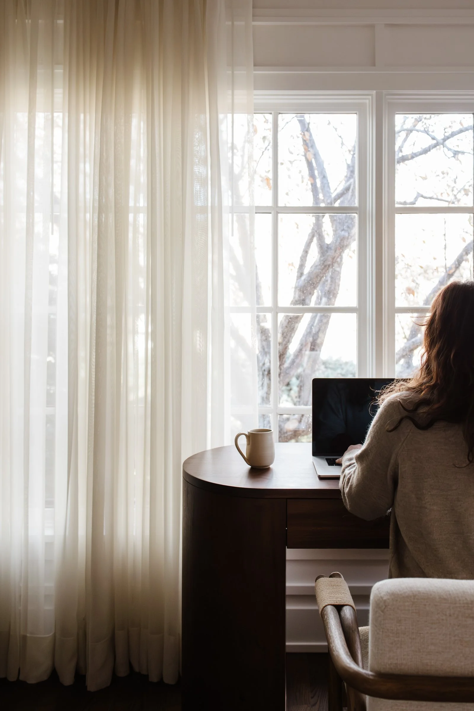 A person sitting at a wooden desk in front of a large window with sheer white curtains, working on a laptop with a mug on the desk.