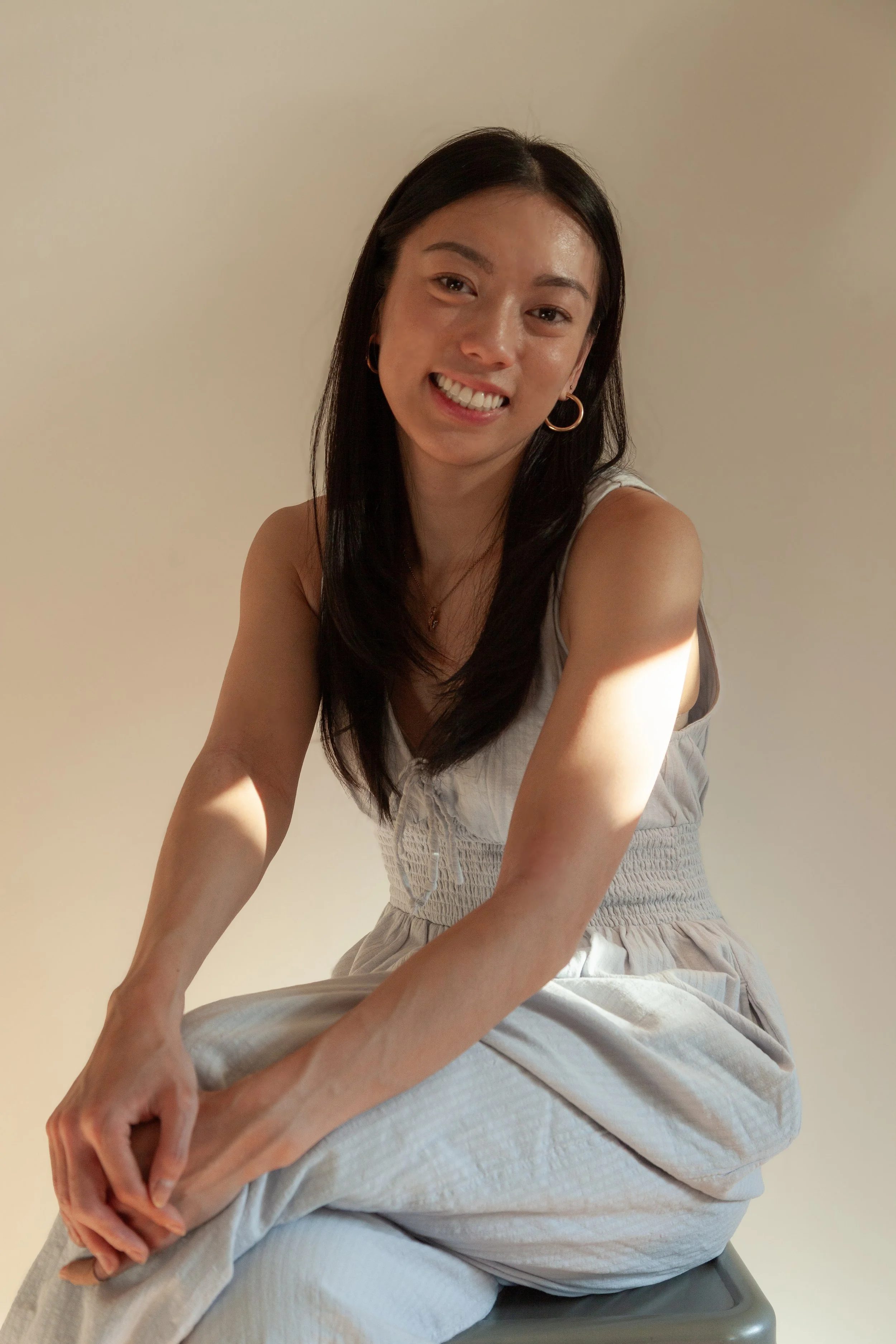 A woman with long dark hair smiling, wearing gold hoop earrings and a light-colored sleeveless dress, sitting on a stool against a plain beige wall.