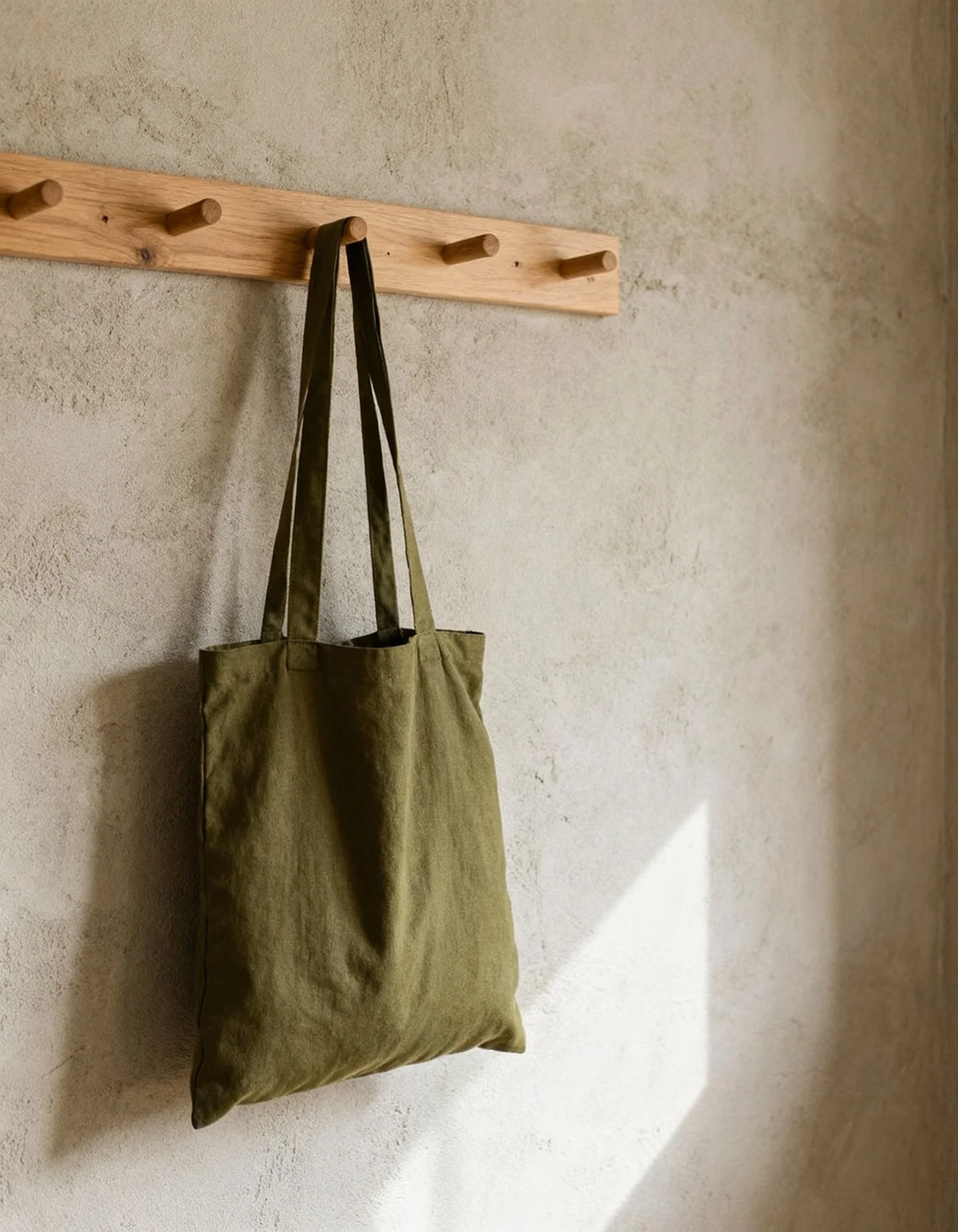Olive green tote bag hanging on a wooden peg on a textured beige wall.